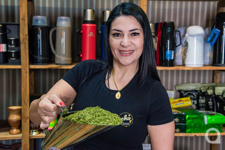 Mulher branca de cabelos pretos sorrindo e segurando uma medida do produto em sua loja especializada em erva-mate em Umuarama